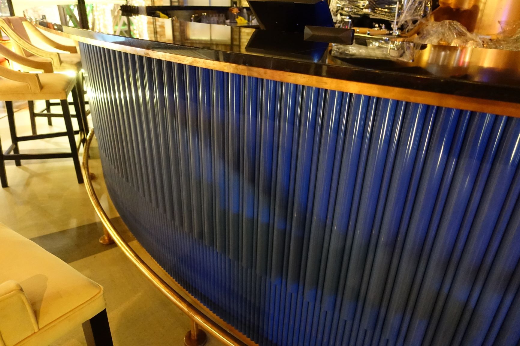 Niebiesko-szara obudowa baru wykonana z pionowych rurek, z czarnym blatem i złotymi wykończeniami, widoczny fragment krzesła barowego.