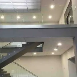 Wnętrze budynku z nowoczesnymi schodami z metalową konstrukcją i szklanymi balustradami na dwóch poziomach, oświetlone lampami przemysłowymi.
