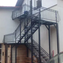 Zewnętrzne metalowe schody z balustradami prowadzące na balkon budynku mieszkalnego, widoczne fragmenty elewacji i samochodu.