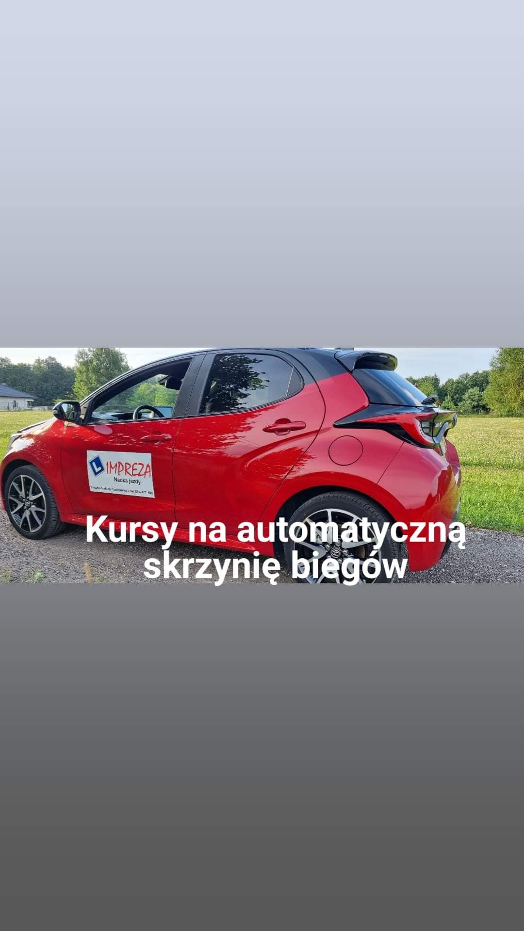 Czerwony samochód z naklejką firmy oferującej kursy na automatyczną skrzynię biegów, zaparkowany na żwirowej drodze z widokiem na zielone pole.