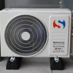 Biała jednostka zewnętrzna klimatyzacji na wspornikach, z widocznym logotypem firmy i danymi kontaktowymi. Montaż przy ścianie z cegły.