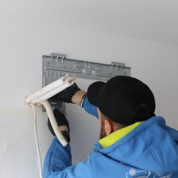 TeslaPV.pl - Fotowoltaika, Klimatyzacja, Pompy Ciepła - Montaż klimatyzacji: instalator w czapce łączy rury do uchwytu na ścianie, zabezpieczonej folią. Widoczne narzędzia i elementy instalacji.