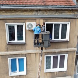 TeslaPV.pl - Fotowoltaika, Klimatyzacja, Pompy Ciepła - Montaż klimatyzatora na elewacji budynku z podnośnika koszowego. Dwóch monterów przy pracy, widok z góry na budynek z oknami i balkonami.