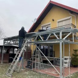 Montaż metalowej konstrukcji zadaszenia tarasu przy żółtym domu z czerwonym dachem, widoczne drabiny i pracownicy w trakcie pracy, na ziemi leży śnieg.