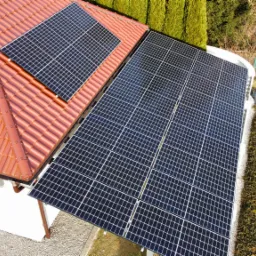 Widok z góry na dom z czerwoną dachówką i panelami słonecznymi, otoczony zielenią.