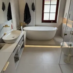 Nowoczesna łazienka w jasnych barwach z wolnostojącą wanną, umywalką nablatową na szafce z szufladami i oświetleniem LED pod wanną.