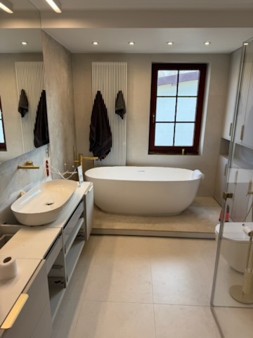 Jasna łazienka z wolnostojącą wanną na podwyższeniu, owalną umywalką nablatową i grzejnikiem drabinkowym w pionowe paski, uzupełniona o złote akcenty w postaci baterii i uchwytów.