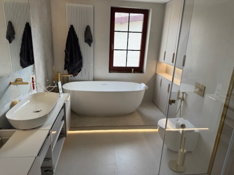 Nowoczesna łazienka w jasnych barwach z wolnostojącą wanną, umywalką nablatową, złotą armaturą i podświetleniem podłogowym.