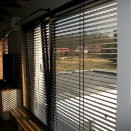 Ciemne drewniane lamele żaluzji poziomej, częściowo zasłonięte, z widokiem na domy i drzewa na zewnątrz.