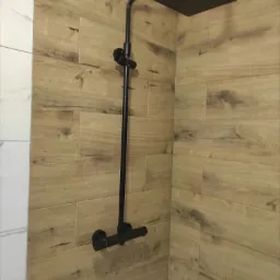 Nowoczesna, czarna deszczownica zamontowana w narożniku kabiny prysznicowej wyłożonej płytkami imitującymi drewno, z fragmentem białych płytek na ścianie po lewej stronie.