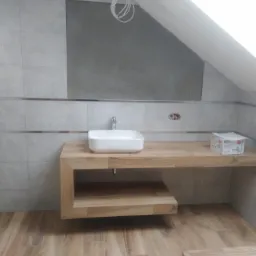 Łazienka w trakcie remontu: biała umywalka nablatowa na drewnianym blacie z półką, szare płytki heksagonalne na ścianach z dekoracyjnym paskiem, surowa ściana pod skosem, podłoga imitująca drewno.