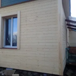 Świeżo wykończona, jasna elewacja drewniana domu z widocznym oknem z białymi ramami i ciemnobrązowym parapetem oraz fragmentem sąsiedniego budynku z zadaszeniem z przezroczystego tworzywa.