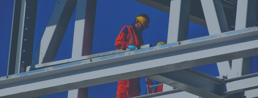 Pracownik w pomarańczowym kombinezonie i żółtym kasku pracuje na metalowej konstrukcji na tle błękitnego nieba, widoczne śruby mocujące elementy konstrukcji.