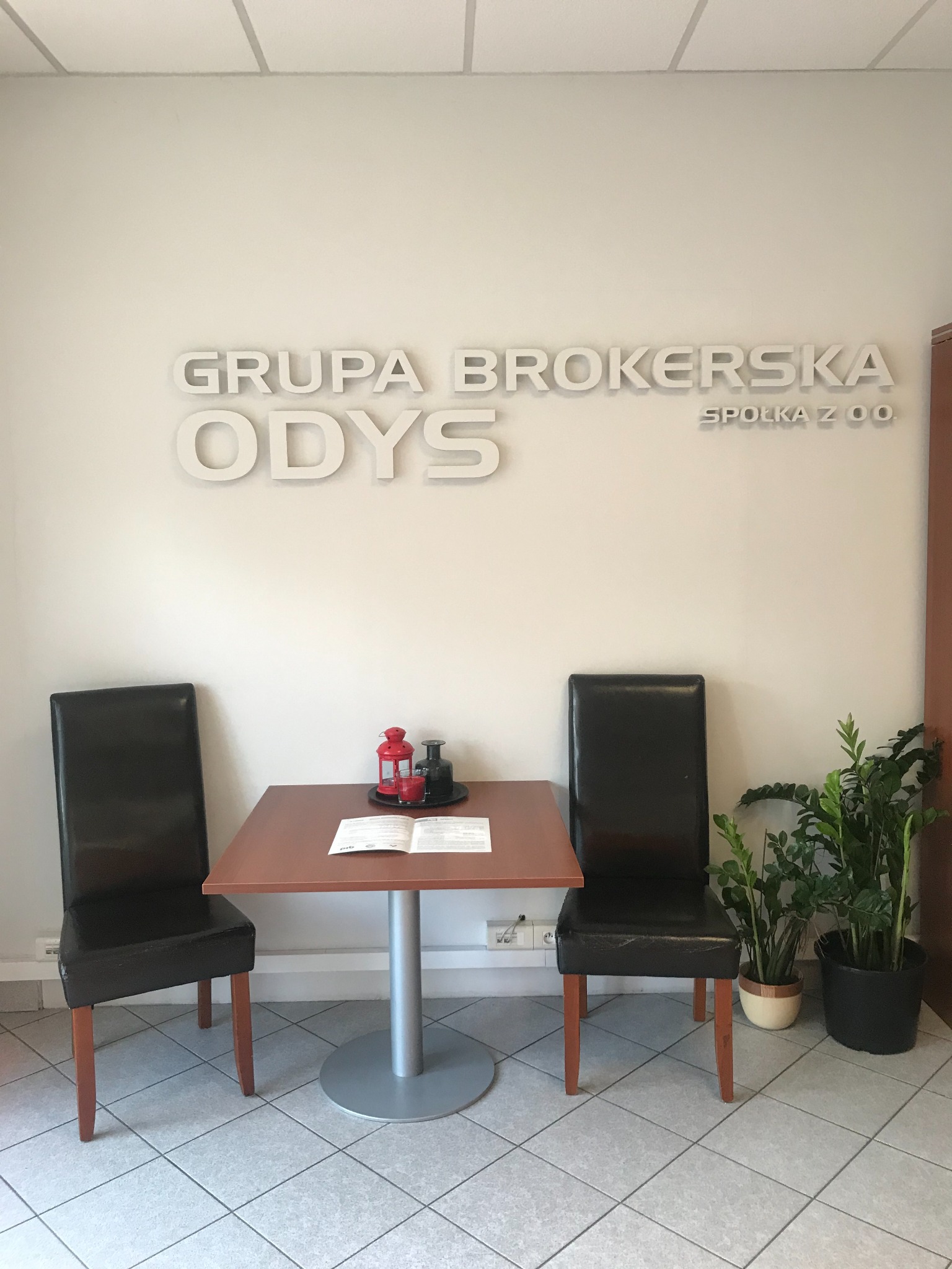 Wnętrze biura Grupy Brokerskiej ODYS Spółka z o.o. z dwoma krzesłami i stolikiem, na którym leżą materiały informacyjne i dekoracje.