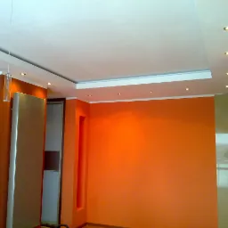 Pomarańczowe ściany w pokoju po malowaniu, z podwieszanym sufitem z oświetleniem punktowym i wiszącymi szklanymi lampami.