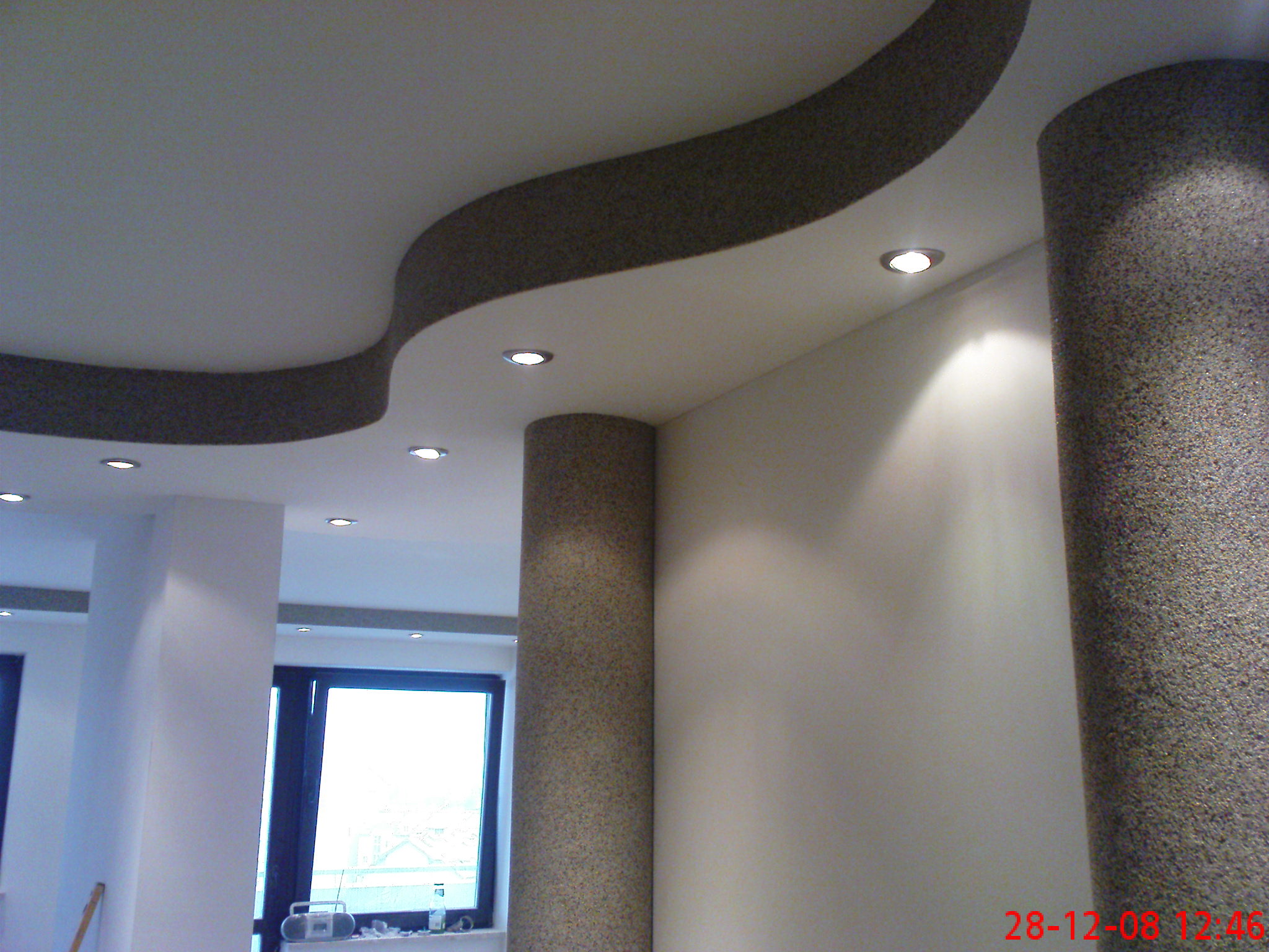 Dekoracyjny sufit podwieszany z falistym wzorem i oświetleniem punktowym, kolumny wykończone tynkiem mozaikowym, widok na okno.