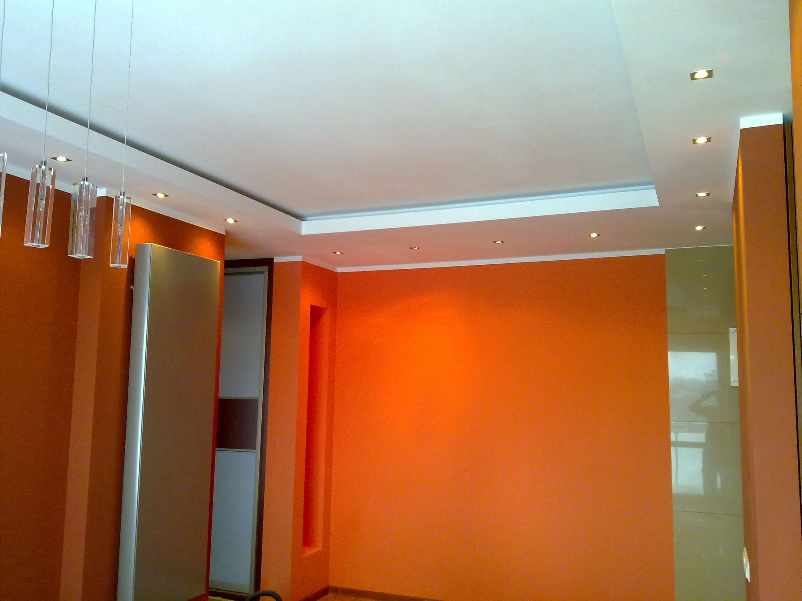 Pomarańczowe ściany w pokoju po malowaniu, z podwieszanym sufitem z oświetleniem punktowym i wiszącymi szklanymi lampami.