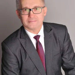 Portret mężczyzny w garniturze w kratę, białej koszuli i bordowym krawacie, na neutralnym szarym tle.