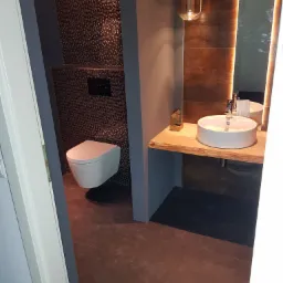 Nowoczesna łazienka z mozaiką na ścianie, podwieszaną toaletą, umywalką nablatową na drewnianym blacie i oświetlonym lustrem.