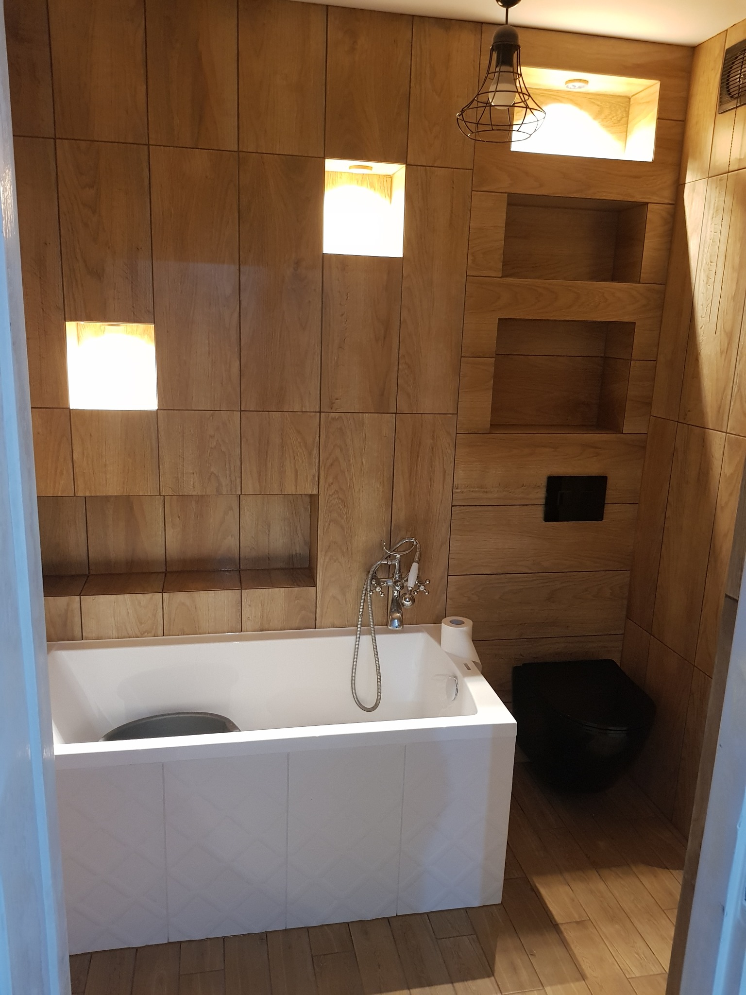 Nowoczesna łazienka wykończona drewnopodobnymi płytkami, z białą wanną z czarną miską, czarną toaletą podwieszaną i oświetleniem punktowym w niszach.