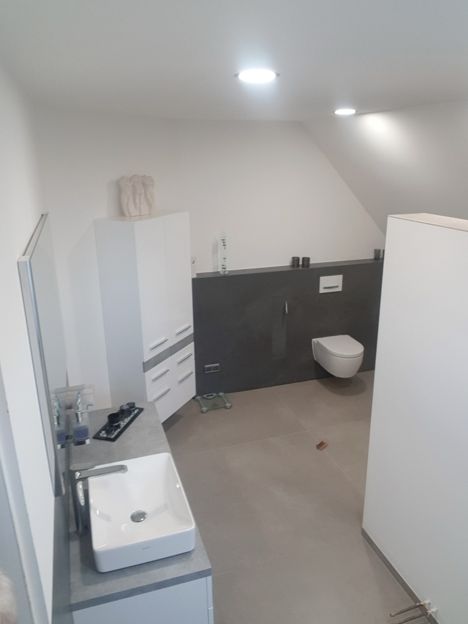 Nowoczesna łazienka na poddaszu z umywalką nablatową, zabudową WC, szarą ścianą i białymi meblami, oświetlona punktowymi lampami sufitowymi.