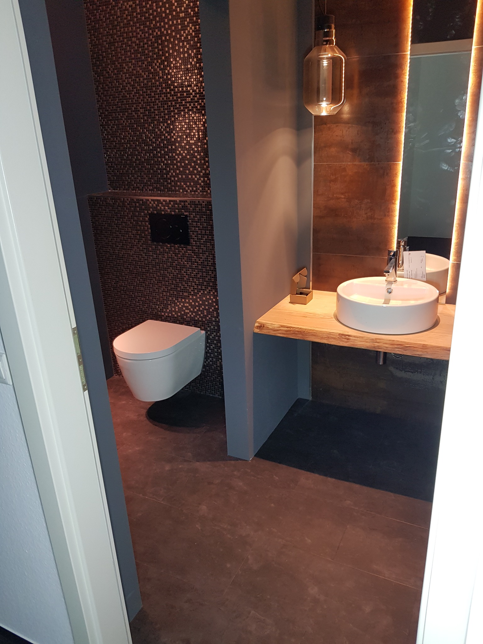 Nowoczesna łazienka z mozaiką na ścianie, podwieszaną toaletą, umywalką nablatową na drewnianym blacie i oświetlonym lustrem.