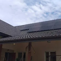 Panel słoneczny na brązowym dachu domu jednorodzinnego z widocznymi rynnami i fragmentem elewacji w kolorze beżowym, na tle błękitnego nieba z chmurami.