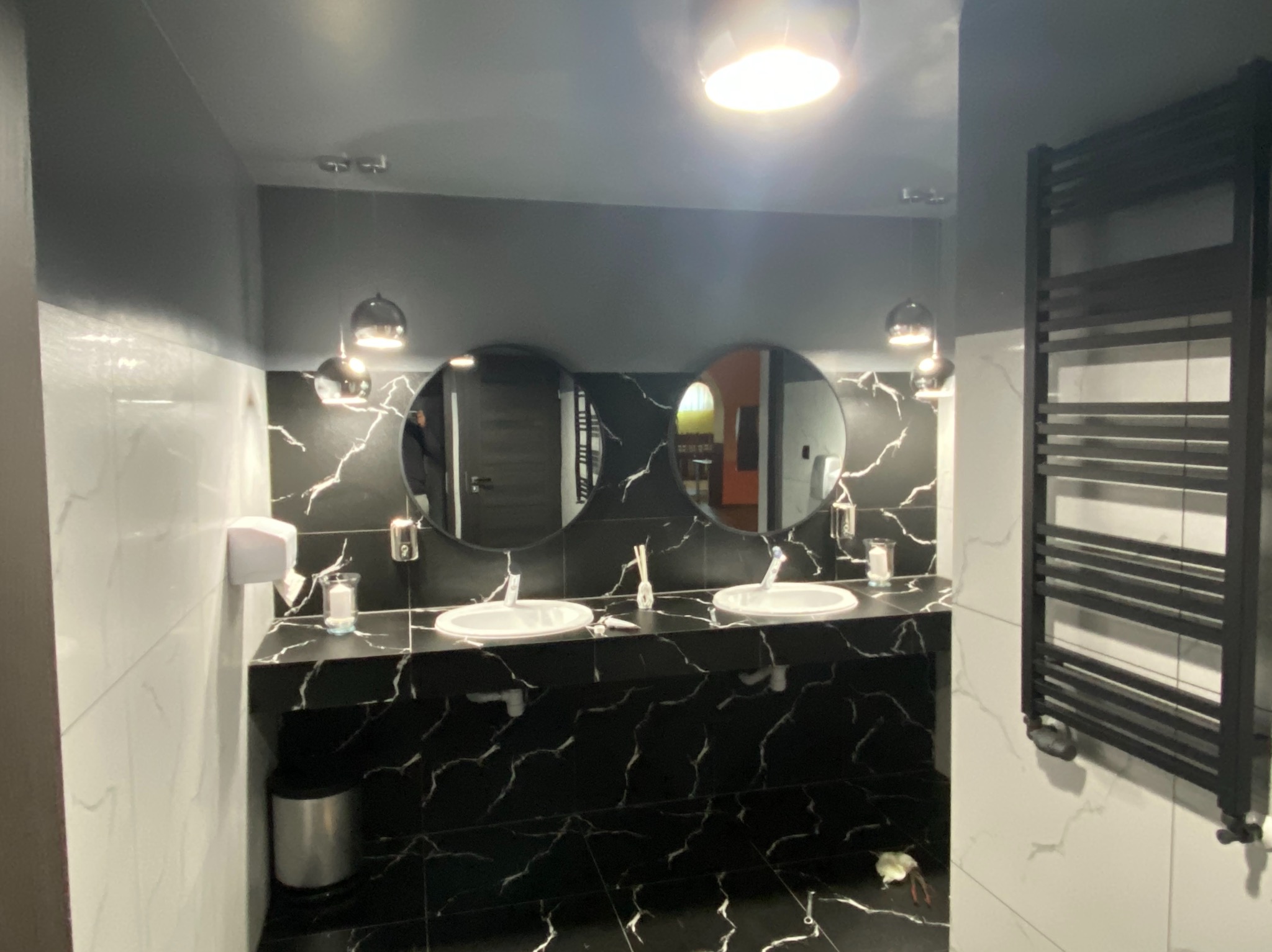 Nowoczesna łazienka z czarnymi blatami i okrągłymi lustrami, białe marmurowe płytki na ścianach, grzejnik drabinkowy w kolorze czarnym, oświetlenie punktowe.