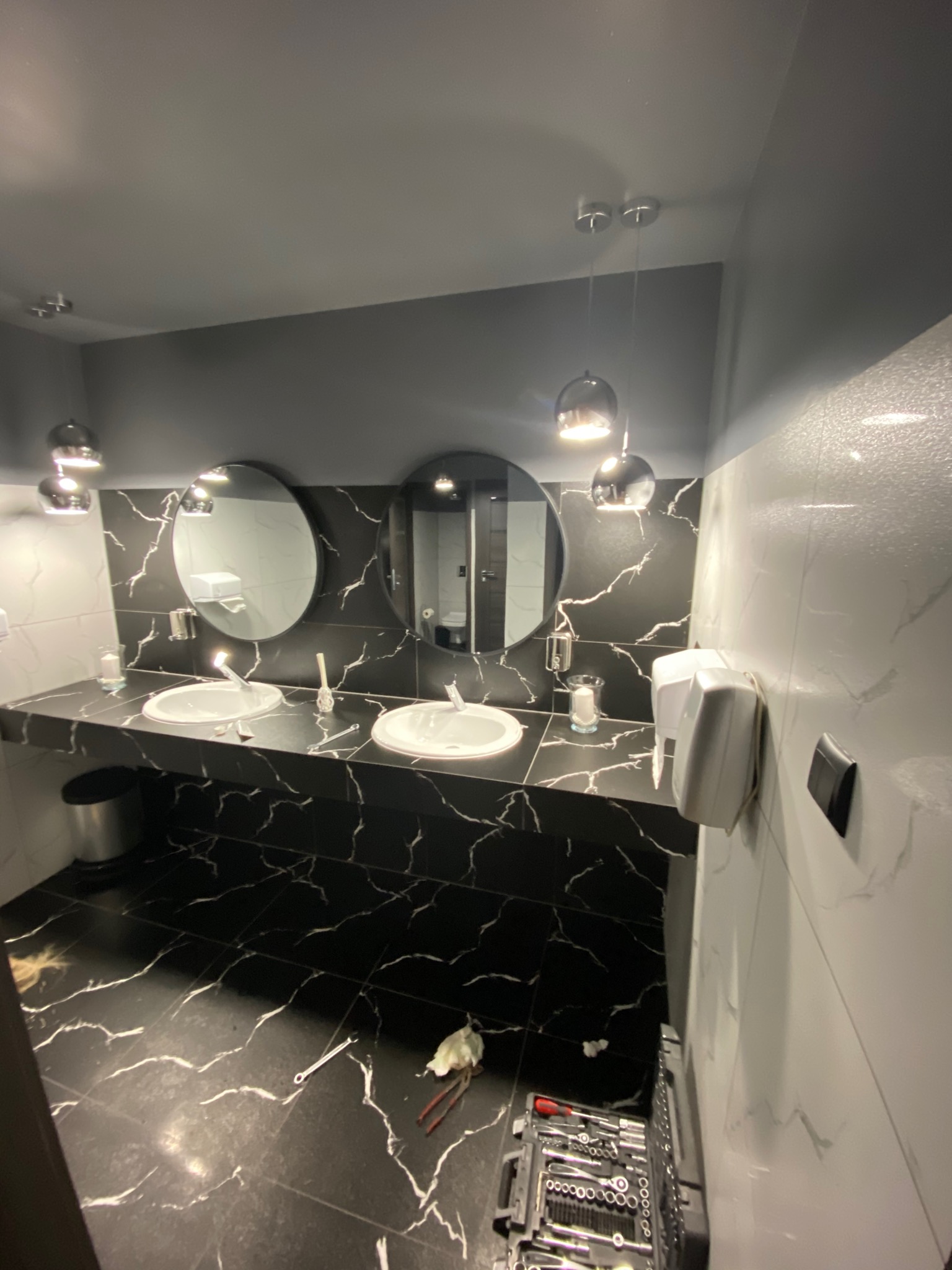 Wykończona łazienka w stylu nowoczesnym z dwoma okrągłymi lustrami, czarnym blatem z białym marmurkowym wzorem i białymi umywalkami. Na podłodze leżą narzędzia i skrzynka z narzędziami, co sugeruje...