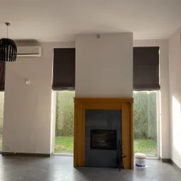 Wnętrze pokoju z szarymi płytkami podłogowymi, drewnianą obudową kominka z szarymi płytkami, dużymi oknami z roletami rzymskimi i wiadrem z farbą w rogu.
