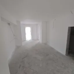 Wnętrze w stanie deweloperskim po pracach przygotowawczych: ściany i sufit pomalowane na biało, podłoga betonowa, widoczne wystające przewody elektryczne, drzwi balkonowe zabezpieczone folią.