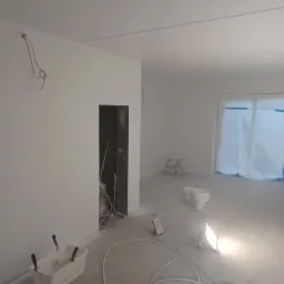 Wnętrze w trakcie odświeżania, ściany pomalowane na biało, widoczne narzędzia malarskie: wiadro z wałkiem, agregat malarski, lampa halogenowa, kable, zabezpieczone folią okno i drzwi.