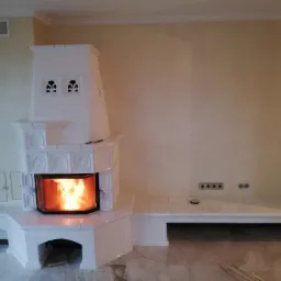 Biały, kaflowy kominek z paleniskiem, wykończony płytkami, z dekoracyjnymi otworami wentylacyjnymi, w jasnym wnętrzu z marmurową podłogą i gładkimi ścianami.