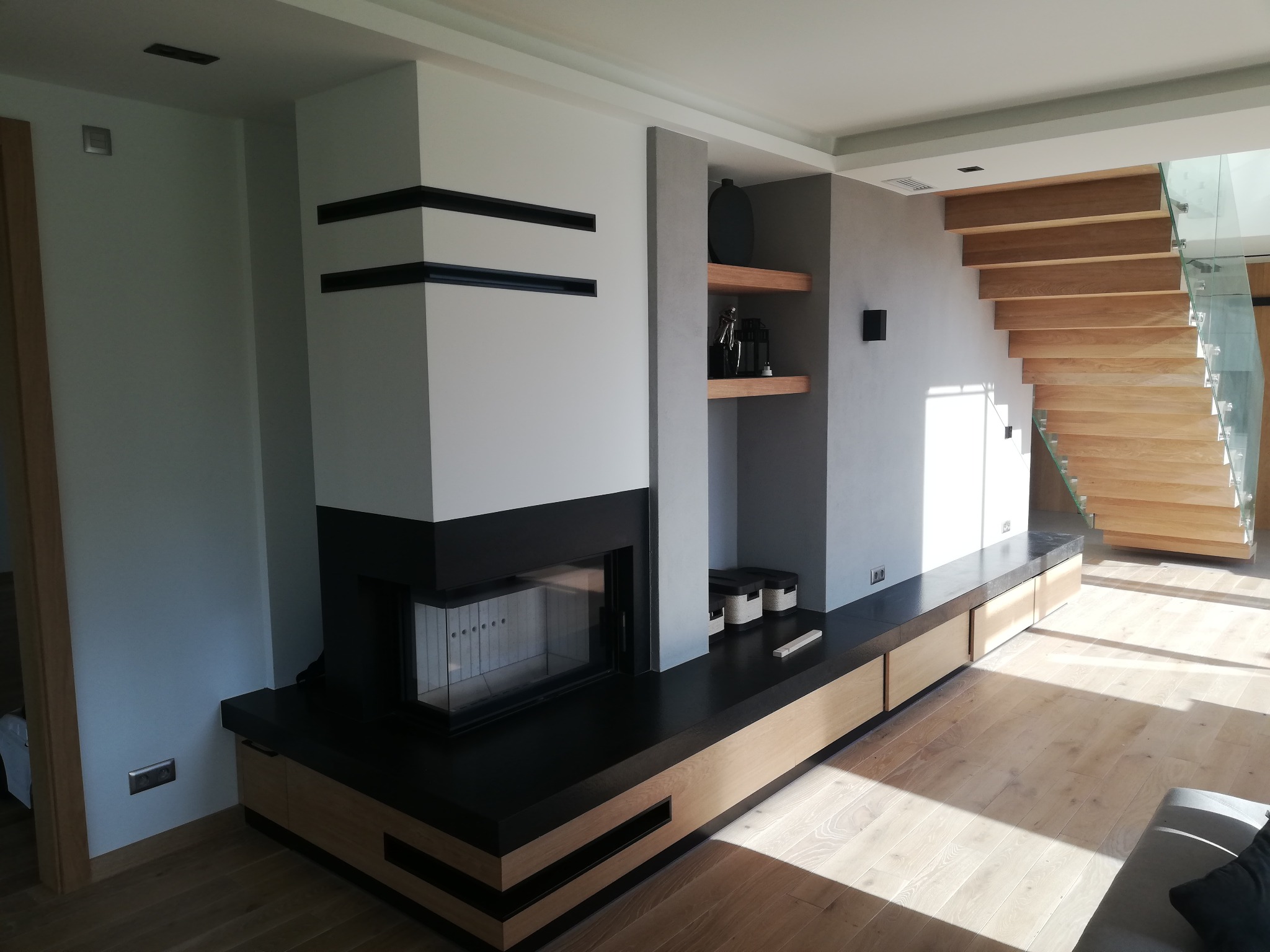 Nowoczesny salon z jasną drewnianą podłogą, białym kominkiem z czarnymi akcentami i drewnianymi schodami z balustradą ze szkła.