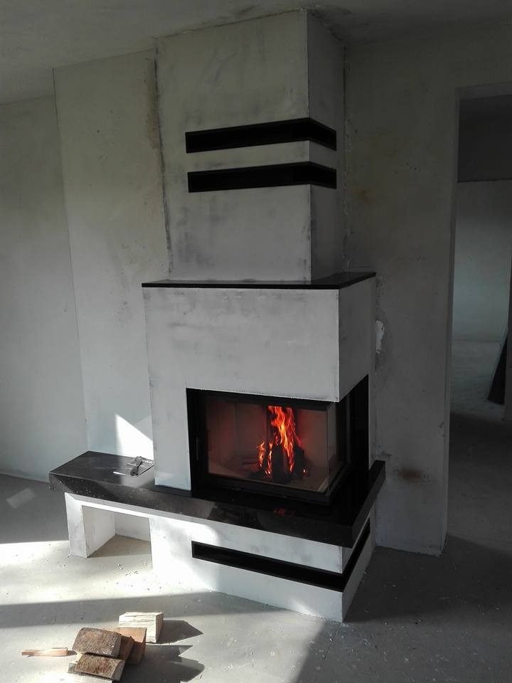 Nowoczesny kominek z czarnymi elementami dekoracyjnymi, widoczne płomienie w palenisku, surowy stan wykończenia wnętrza.