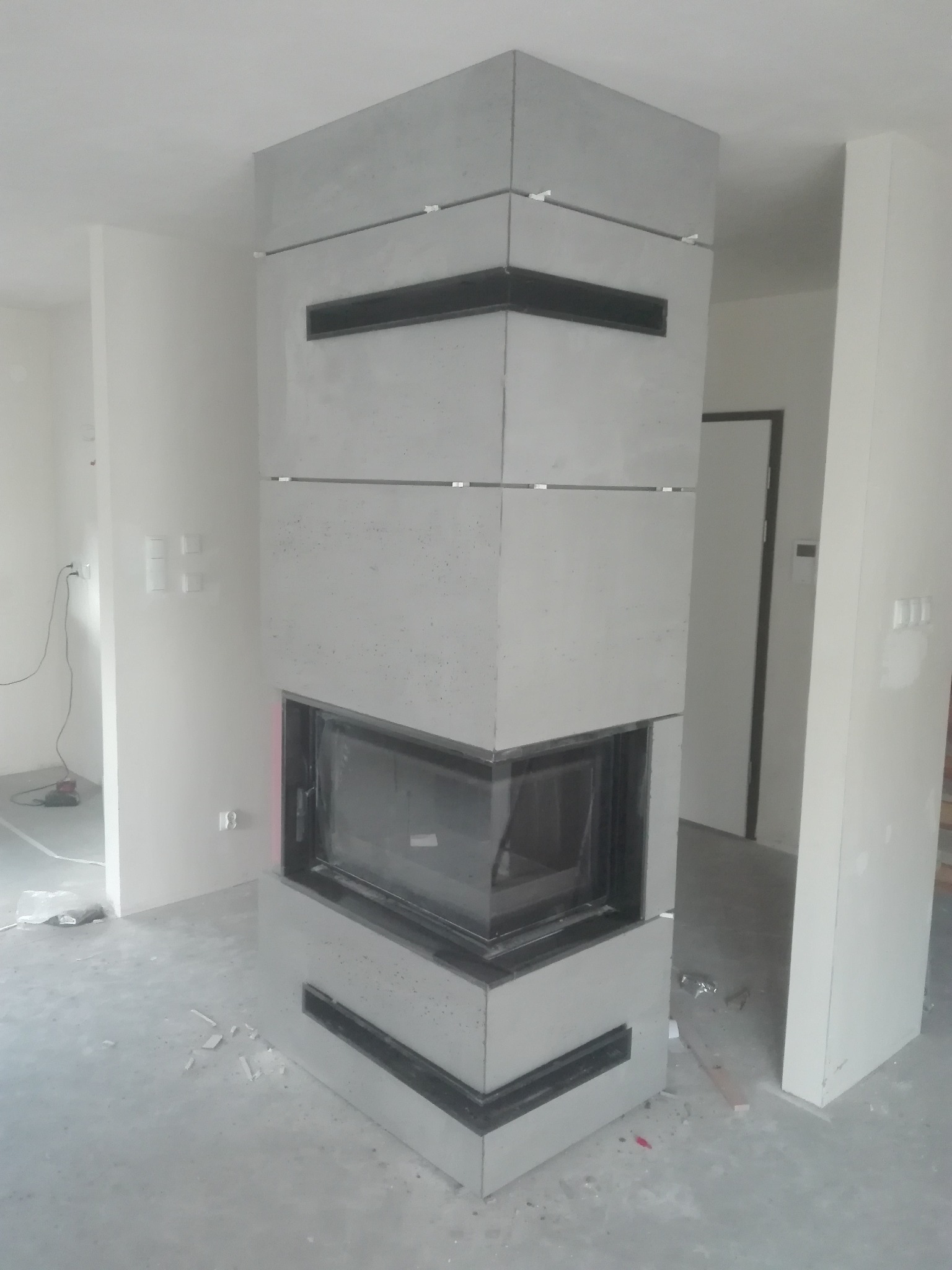 Kanciasty kominek obłożony szarymi płytami imitującymi beton, z czarnymi wstawkami, w surowym wnętrzu w trakcie wykończenia, widoczne białe ściany i niedokończona podłoga.