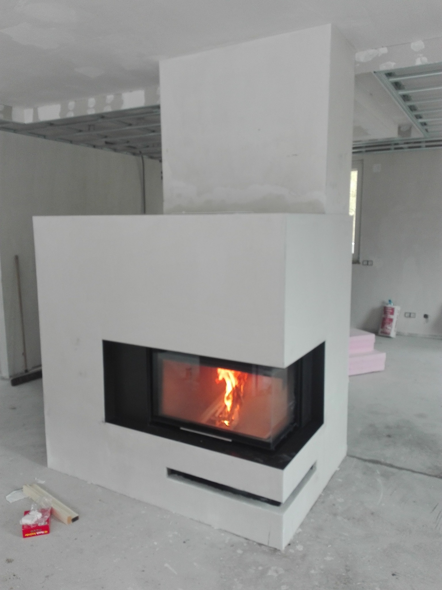 Nowoczesny, biały kominek z czarnym obramowaniem w surowym wnętrzu, z widocznym płomieniem, w trakcie prac wykończeniowych, widoczne materiały budowlane i konstrukcja sufitu podwieszanego.