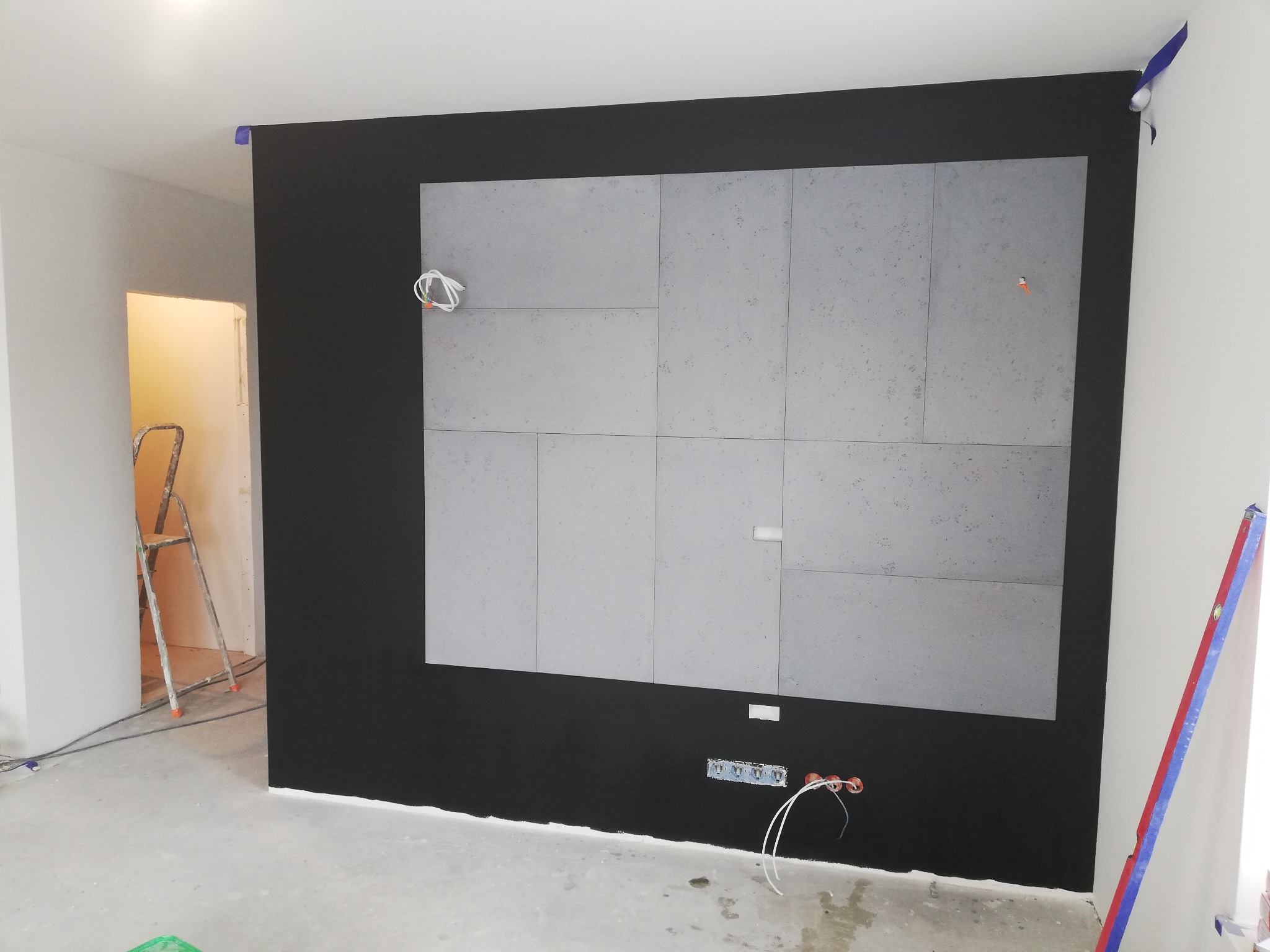 Ściana w trakcie wykańczania z szarymi płytami imitującymi beton na czarnym tle, widoczne przewody elektryczne i poziomica przy ścianie, drabina w tle.