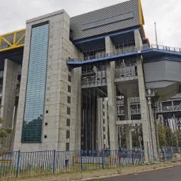 Industrialna konstrukcja podnośni z betonu i stali, widoczne elementy instalacji energetycznych, żółte i niebieskie akcenty kolorystyczne, szara fasada budynku z pionowym oknem.