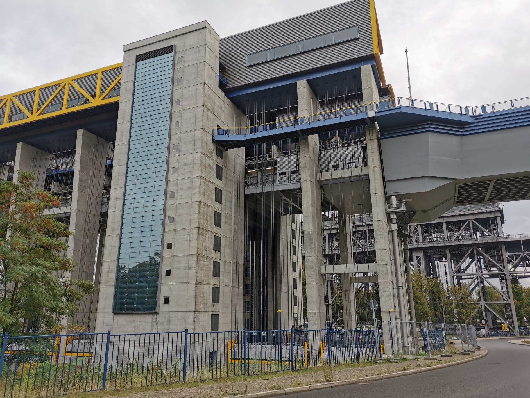 Industrialna konstrukcja podnośni z betonu i stali, widoczne elementy instalacji energetycznych, żółte i niebieskie akcenty kolorystyczne, szara fasada budynku z pionowym oknem.