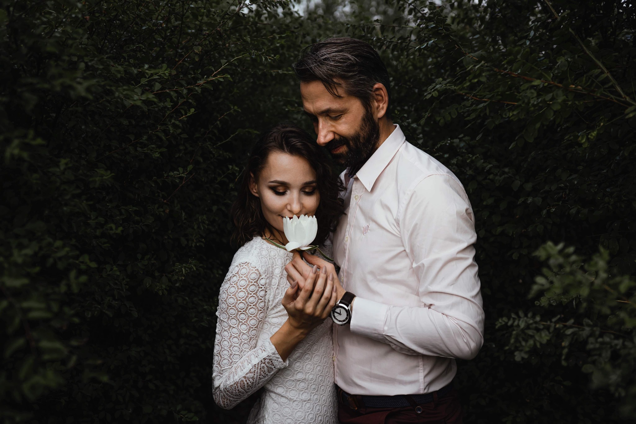 Para zakochanych: kobieta w koronkowej sukience wącha białego tulipana, mężczyzna w różowej koszuli obejmuje ją czule, tle zielone krzewy.