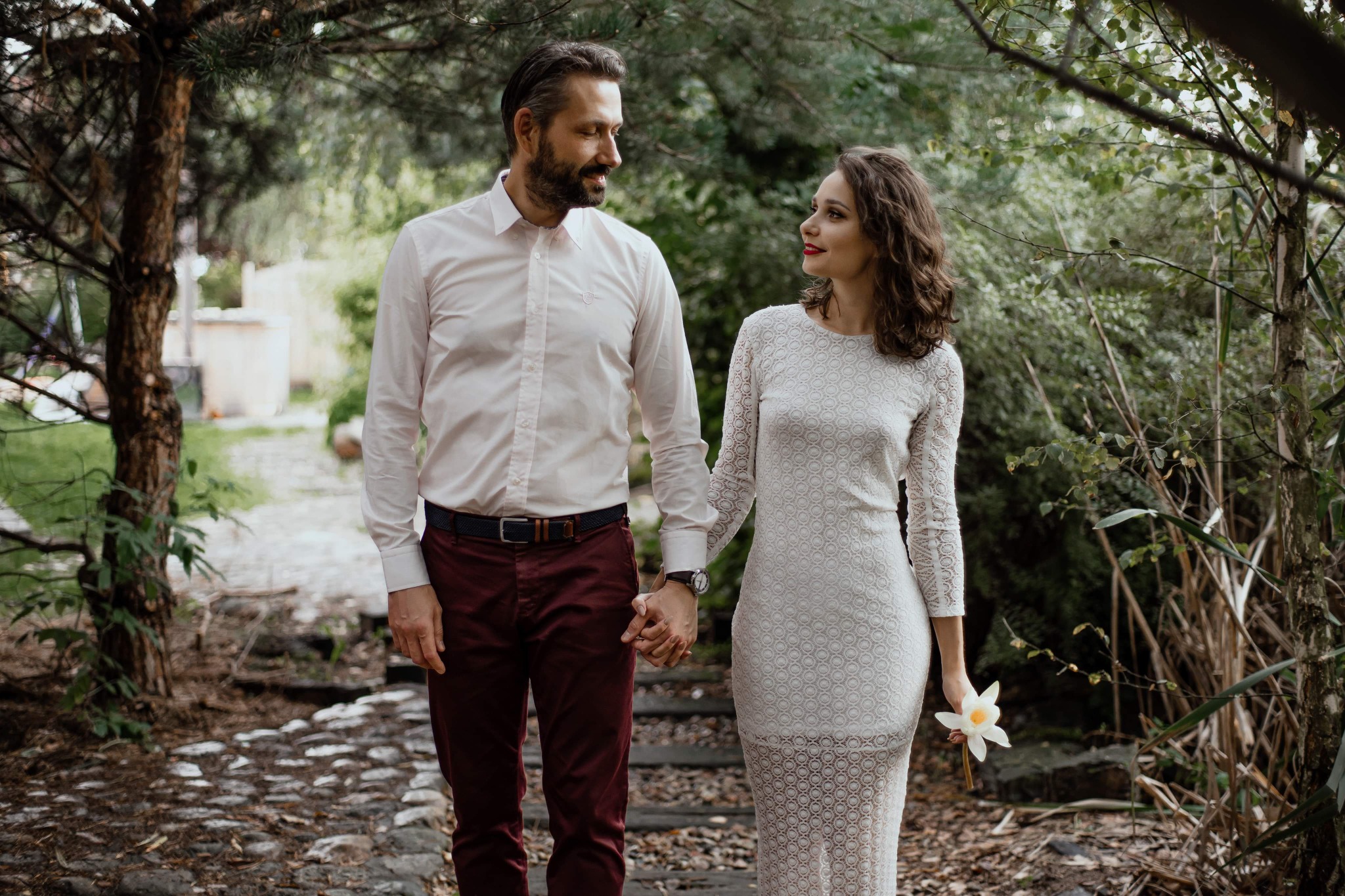 Para zakochanych idzie ścieżką w lesie, trzymając się za ręce; kobieta w koronkowej sukience trzyma biały kwiat.