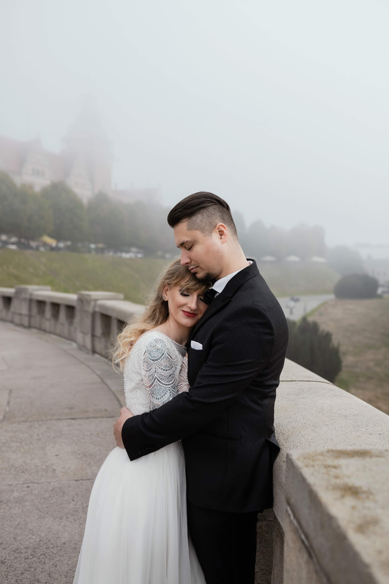 Para młoda obejmująca się na tle zamglonego krajobrazu miejskiego, panna młoda w koronkowej sukni, pan młody w czarnym garniturze.