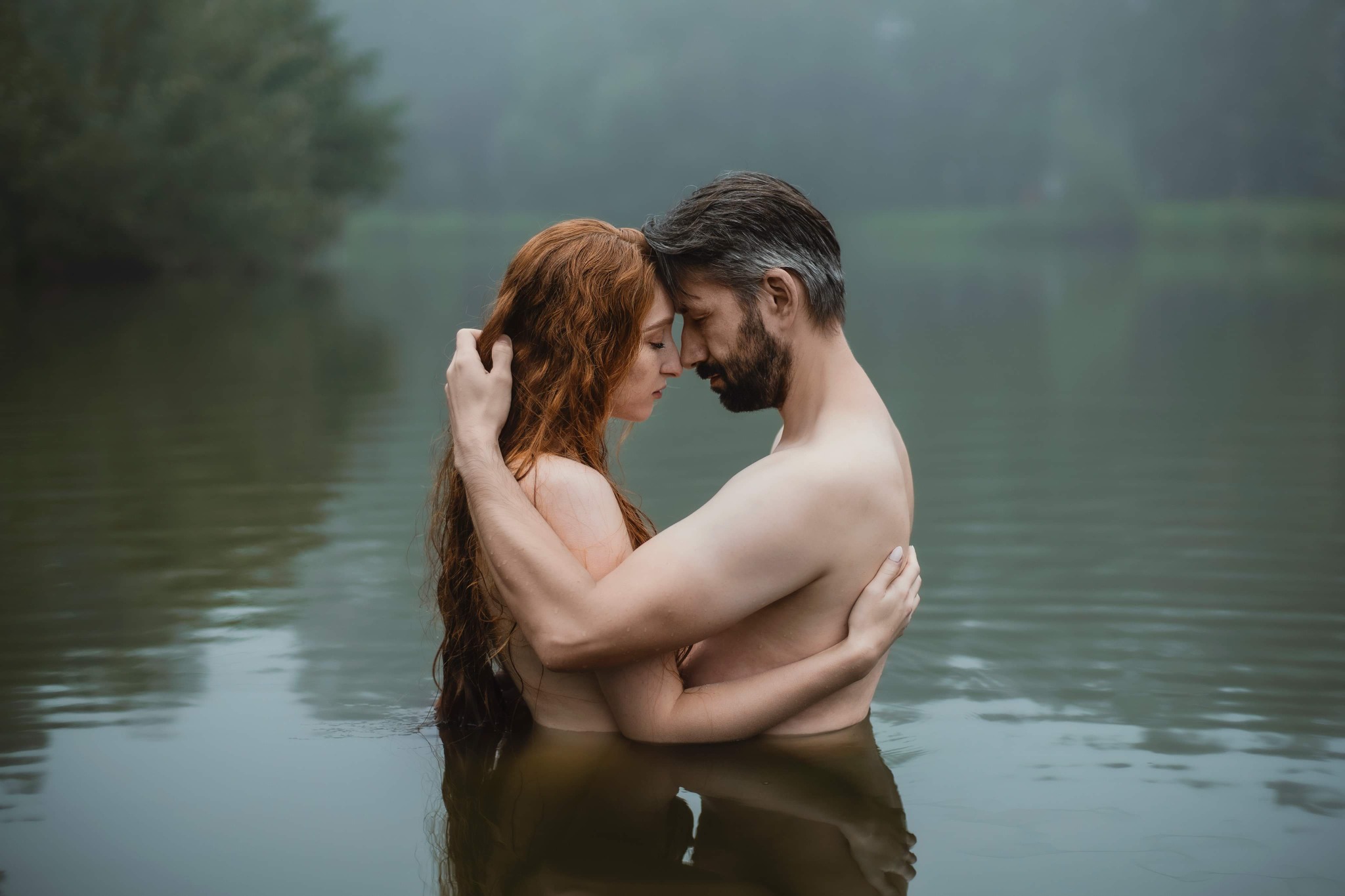 Zakochana para w wodzie, kobieta z rudymi włosami obejmowana przez mężczyznę z brodą, oboje stoją zanurzeni w jeziorze, w tle mglisty krajobraz.