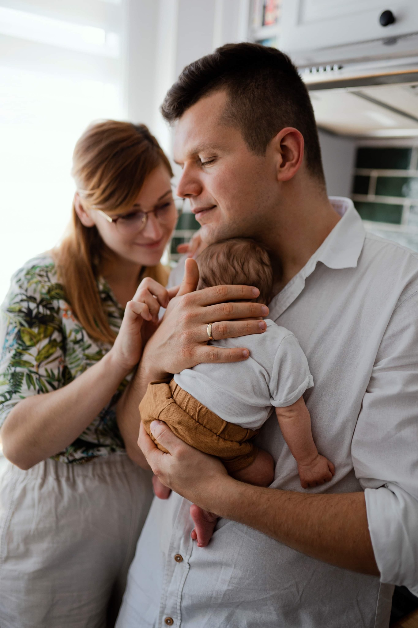 Ojciec trzyma noworodka w ramionach, matka patrzy z czułością, delikatne światło w tle kuchni z zieloną mozaiką.