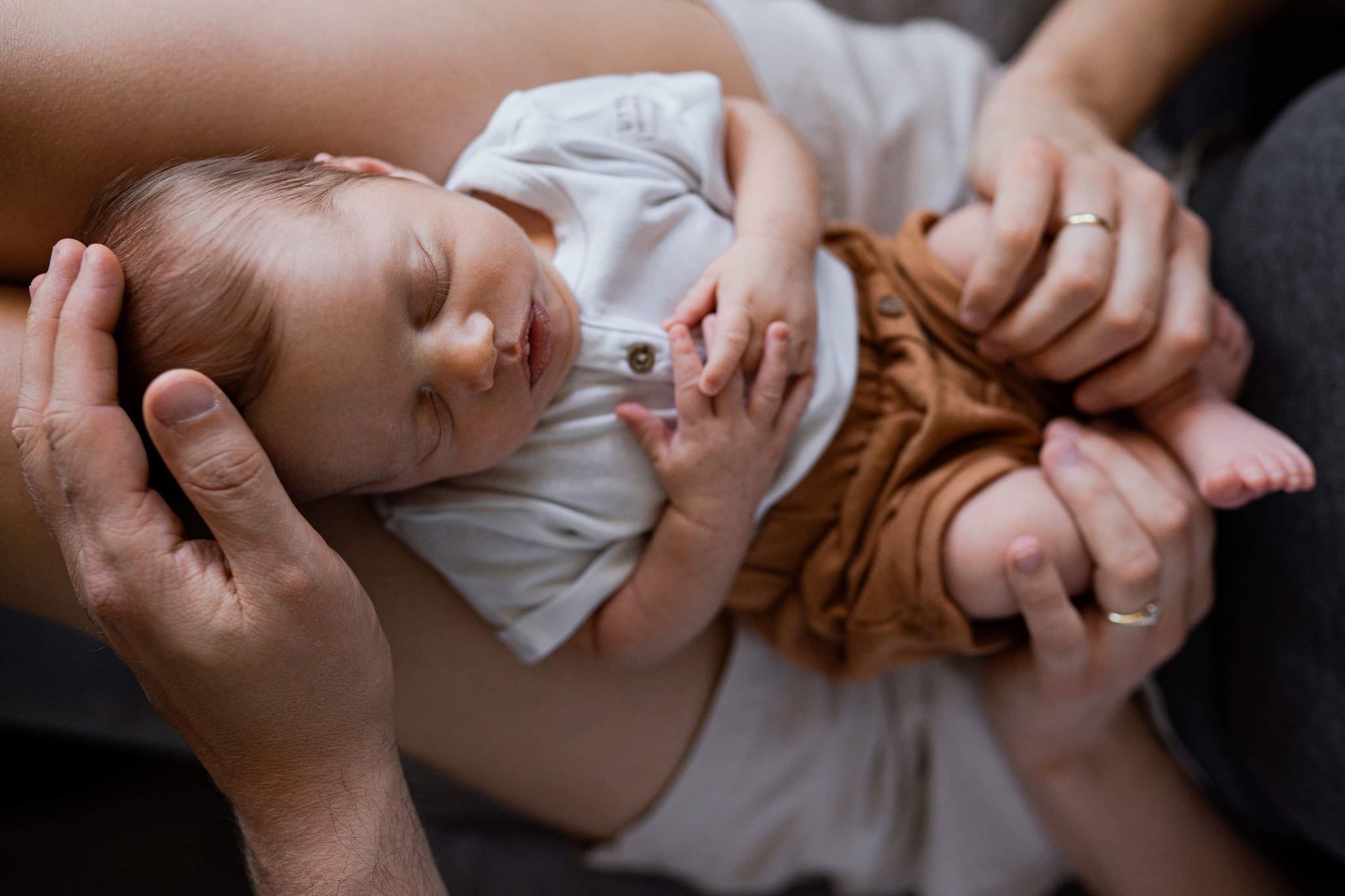 Śpiący noworodek w beżowym ubranku, trzymany przez dwie osoby z obrączkami na palcach, delikatne oświetlenie, bliski kadr.