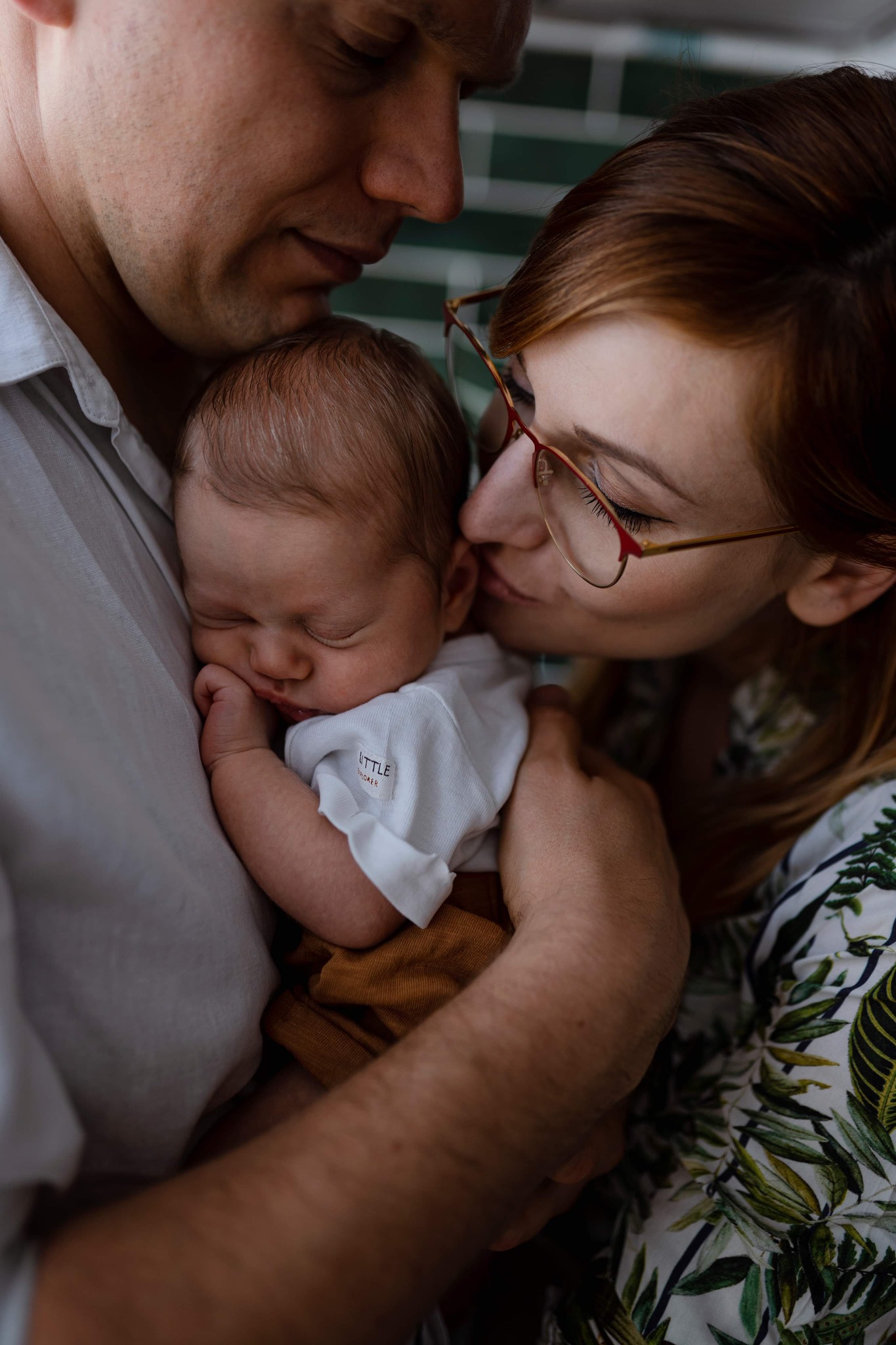 Rodzice tulą śpiącego noworodka: tata trzyma dziecko, mama całuje w policzek, oboje patrzą z miłością.