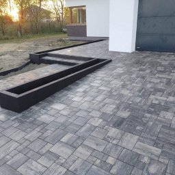 Totalbruk Wojciech Delegacz - Nowoczesna nawierzchnia z szarej kostki brukowej ułożonej we wzór, z czarnymi obrzeżami i elementami dekoracyjnymi, w tle widoczny fragment domu z bramą garażową.