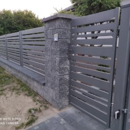 Totalbruk Wojciech Delegacz - Nowoczesne ogrodzenie panelowe w kolorze grafitowym z poziomymi przęsłami, słupki z szarej kostki łupanej, furtka z domofonem, widok z zewnątrz.