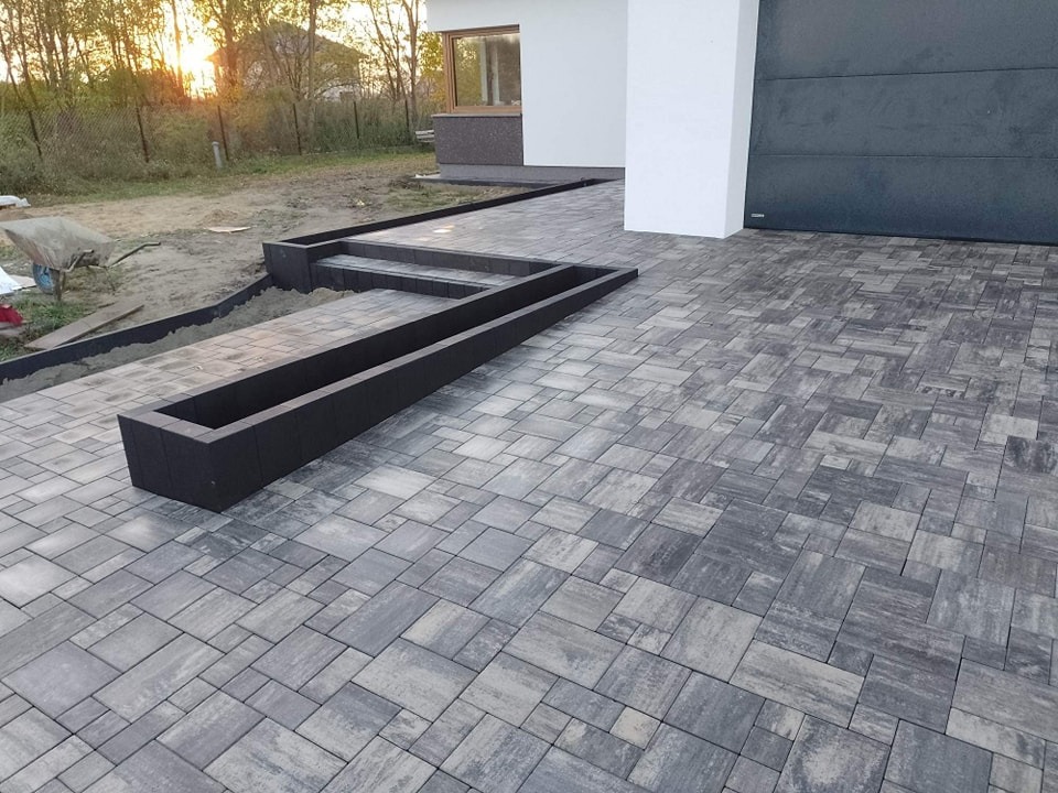 Nowoczesna nawierzchnia z szarej kostki brukowej ułożonej we wzór, z czarnymi obrzeżami i elementami dekoracyjnymi, w tle widoczny fragment domu z bramą garażową.